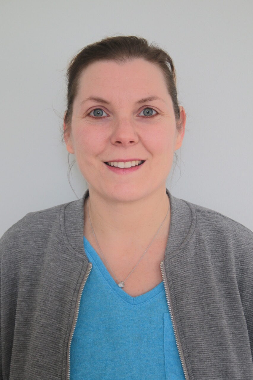 Eine Frau mit heller Haut und braunem, zurückgestecktem Haar lächelt in die Kamera. Sie trägt einen grauen Pullover mit Reißverschluss über einem blauen Oberteil und eine silberne Halskette und steht vor einem schlichten hellen Hintergrund.