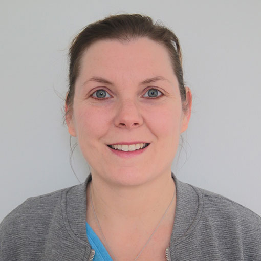 Eine Frau mit heller Haut, braunem zurückgebundenem Haar und blauen Augen lächelt in die Kamera. Sie trägt eine graue Jacke über einem blauen Oberteil und steht vor einem schlichten hellen Hintergrund.