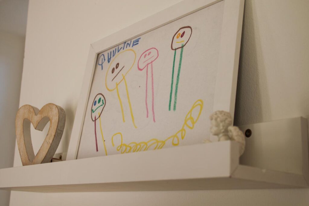 Auf einem weißen Regal stehen eine gerahmte Kinderzeichnung mit fünf bunten Strichmännchen, ein hölzernes Herz und eine kleine weiße Engelsfigur - eine warme Note in dieser Praxis von Logopädie Gummersbach. Die Zeichnung zeigt Gesichter und Linien in hellen Farbtönen.