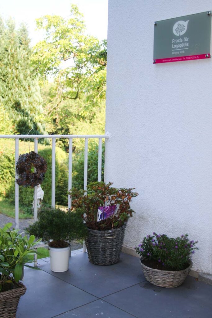 Topfpflanzen und ein Blumenkorb schmücken einen gefliesten Balkon neben einem weißen Geländer. An der Wand ist ein grünes und rosafarbenes Schild von Logopädie Gummersbach angebracht, und im Hintergrund sind grüne Bäume zu sehen.