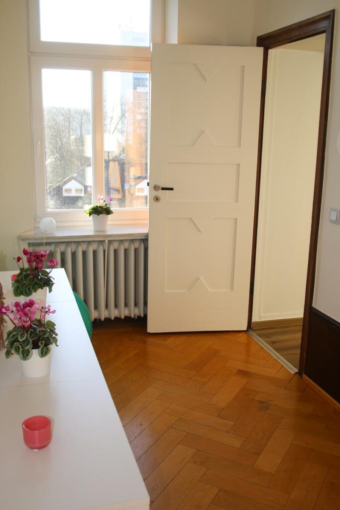 Ein heller Raum mit einem hölzernen Fischgrätenboden, einem weißen Tisch mit rosafarbenen Topfblumen - perfekt für die Sitzungen von Logopädie Gummersbach -, einem Heizkörper unter einem großen Fenster und einer offenen weißen Tür, die in einen anderen Raum führt.