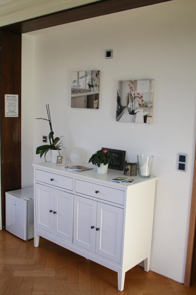 Ein weißes Sideboard mit Schubladen und Schränken steht bei Logopädie Gummersbach an einer Wand. Darauf befinden sich eine getopfte Orchidee, eine Glaskugel, eine kleine Kreidetafel und eine Kerze. An der Wand darüber hängen zwei gerahmte Fotos.