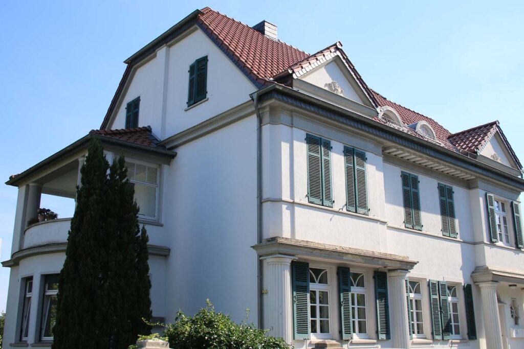 Ein großes zweistöckiges weißes Haus mit grünen Fensterläden und rotem Ziegeldach, das die Logopädie Gummersbach beherbergt. Das Gebäude hat mehrere Fenster, eine kleine überdachte Veranda und einen Garten mit Bäumen und Sträuchern vor dem Haus.