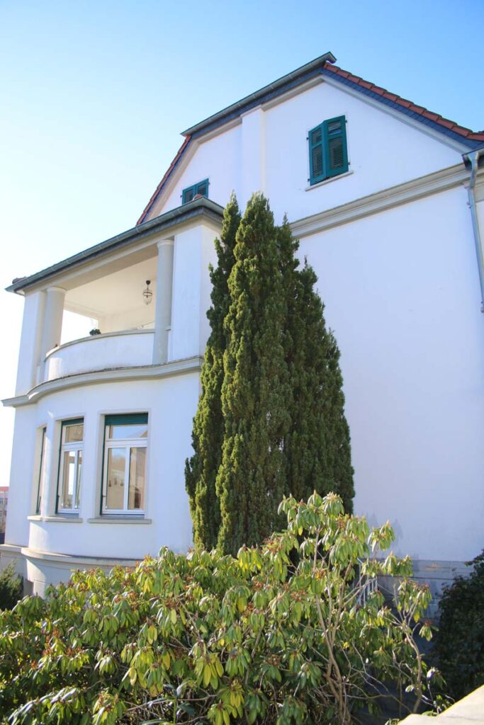 Ein hoher, grüner Baum steht vor einem weißen, zweistöckigen Haus mit grünen Fensterläden und einem Balkon - dem Zuhause von Logopädie Gummersbach. Im Vordergrund sind Sträucher zu sehen, und der Himmel ist klar und blau.