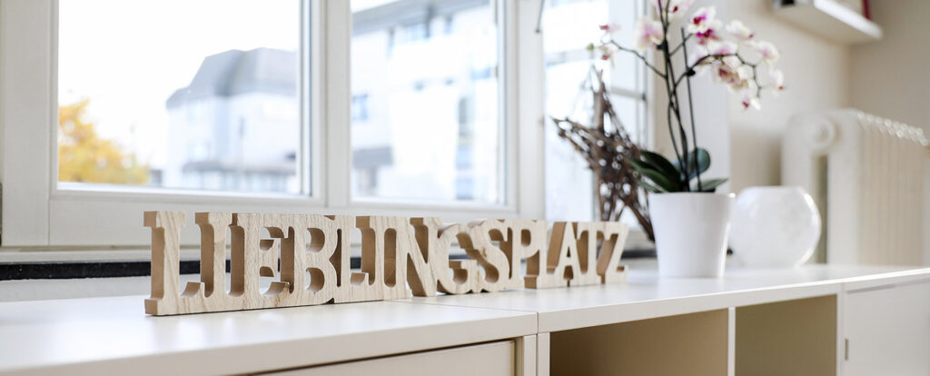 Holzbuchstaben mit dem Schriftzug "LIEBLINGSPLATZ" auf einem weißen Schrank in der Nähe eines Fensters der Logopädie Gummersbach, mit einer getopften Orchidee und einer weißen Vase im Hintergrund; draußen sind Wohnhäuser zu sehen.