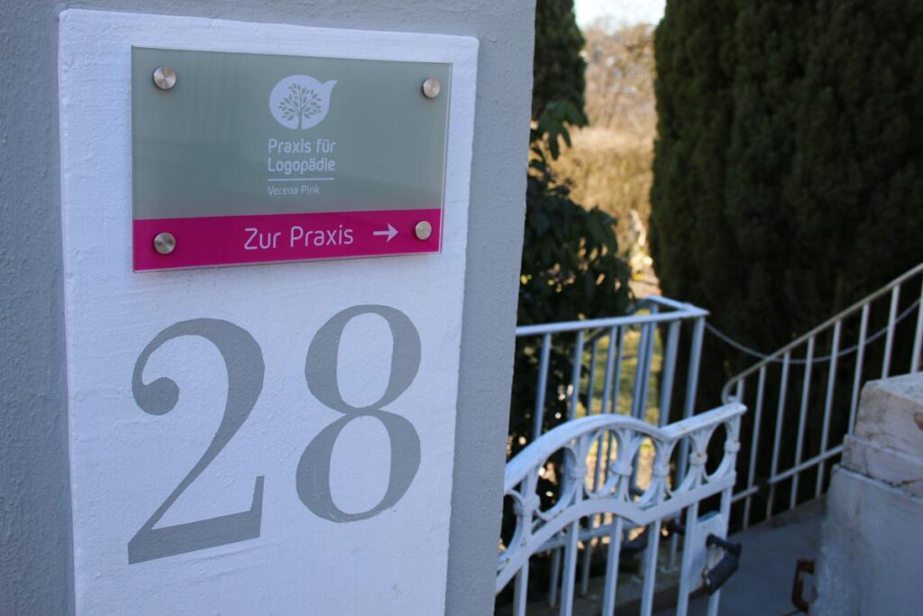 Auf einem Schild an einer weißen Wand steht "Praxis für Logopädie, Verena Pink" mit einem Pfeil und "Zur Praxis". Darunter steht die Nummer 28. Der Eingang zur Praxis für Logopädie Gummersbach ist durch ein weißes Tor, eine Treppe und die umgebende Grünanlage gekennzeichnet.