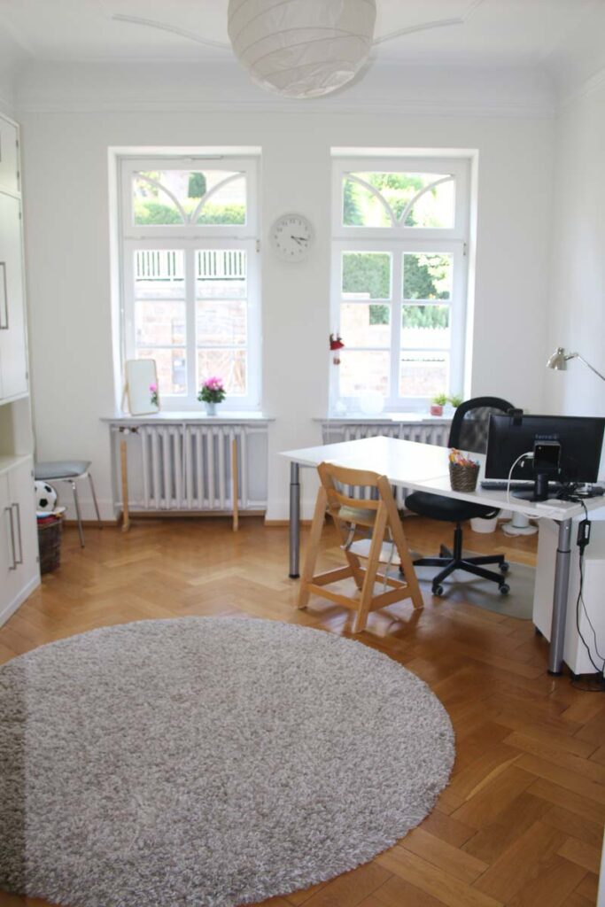 Helles Home-Office mit Holzboden, rundem beigen Teppich, weißem Schreibtisch mit Computer, schwarzem Stuhl, Holzstuhl, Topfpflanzen und großen Fenstern, die natürliches Licht hereinlassen - ein idealer Raum für Logopädie Gummersbach-Sitzungen oder Fernarbeit.