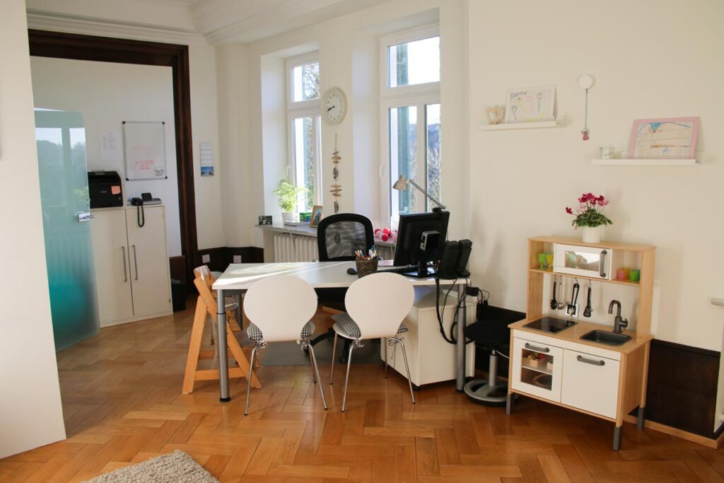 Ein helles Büro von Logopädie Gummersbach mit einem Schreibtisch, einem Computer und zwei weißen Stühlen. Eine Kinderspielküche und Spielzeug füllen die Ecke, während das Sonnenlicht durch große Fenster auf Holzböden und eine gepflegte Einrichtung fällt.