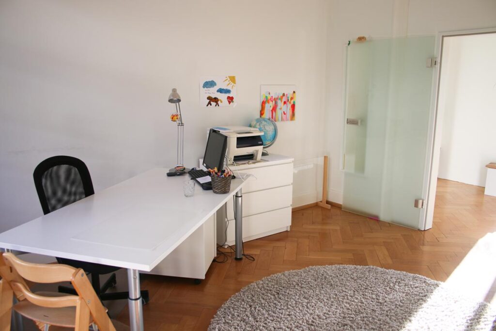 Ein helles, aufgeräumtes Büro für Logopädie Gummersbach mit weißem Schreibtisch, Stuhl, Drucker, Globus und Kunstzubehör. Die Kunstwerke der Kinder hängen über einem weißen Schrank. Ein runder Teppich bedeckt einen Teil des Holzbodens neben einer Glastür.
