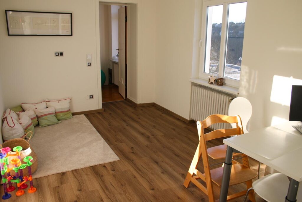 Heller Raum mit Holzboden, gemütlicher Spielecke mit Spielzeug und Kissen auf einem Teppich, weißem Schreibtisch und Stuhl, Holzhochstuhl, sonnigem Fenster und einem Durchgang - perfekt für Logopädie Gummersbach Sitzungen in einladender Atmosphäre.